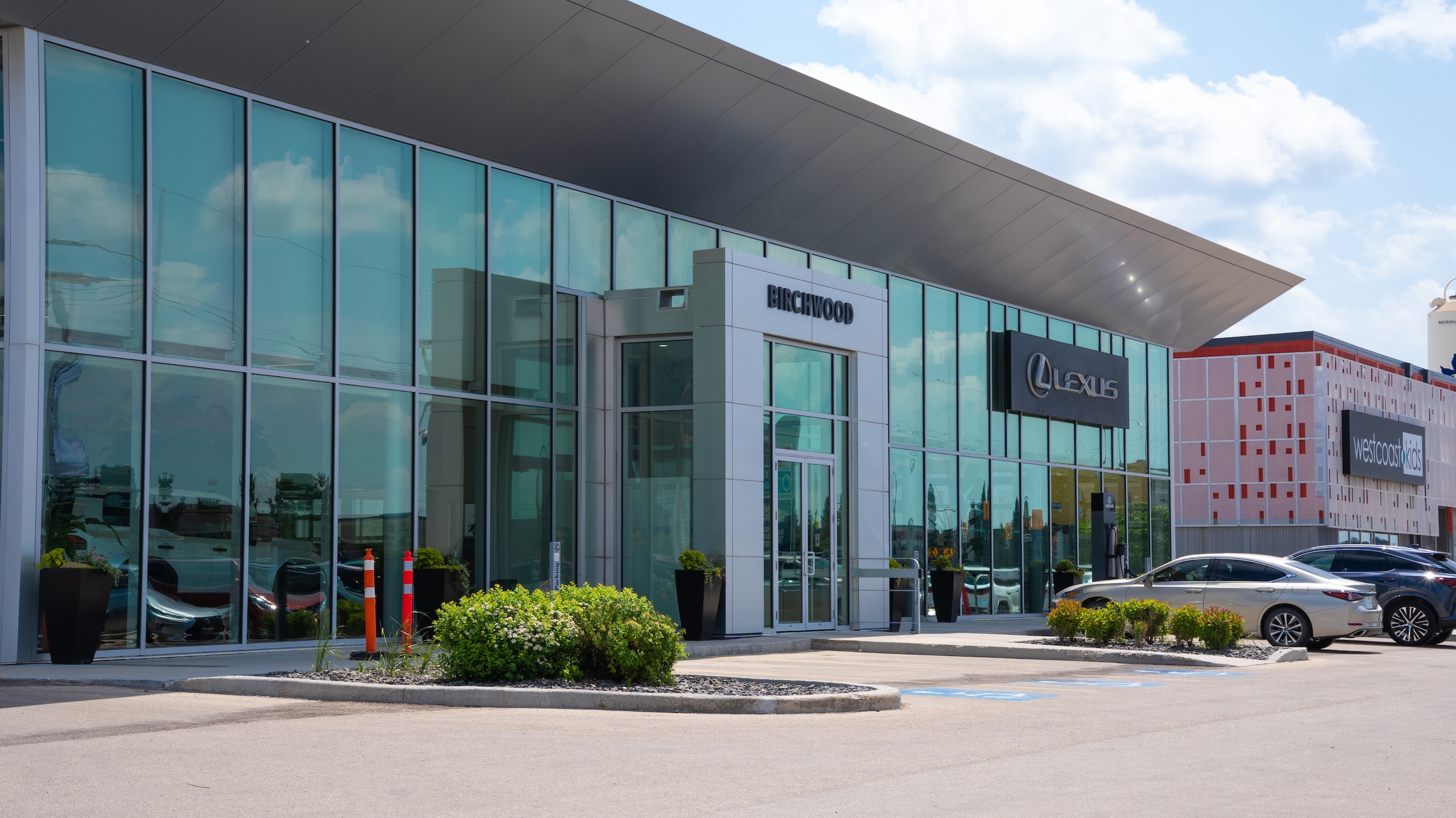 Exterior of Birchwood Lexus dealership featuring sleek glass architecture, the Lexus logo, and adjacent storefronts, including West Coast Kids, under a bright sky.